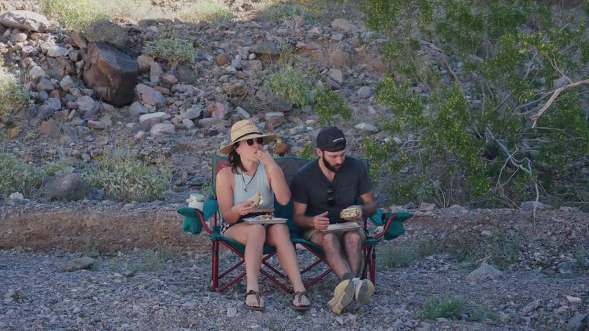 A couple on an outdoor adventure takes a break to eat lunch together on a camping chair in a rocky desert wash