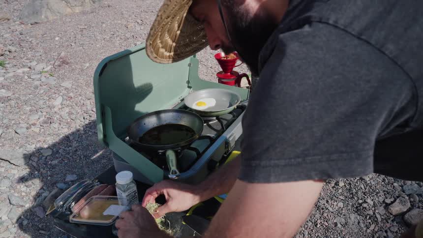 Man in a straw hat cooks a gourmet breakfast sandwich with eggs and avocado on a portable camp stove in a rustic outdoor setting