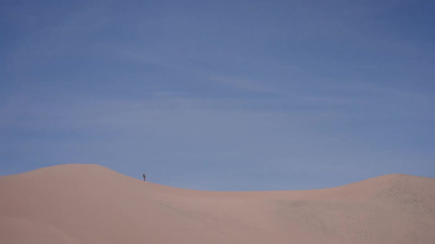 A lone person walks along the ridge of a massive sand dune under a vast, clear blue sky