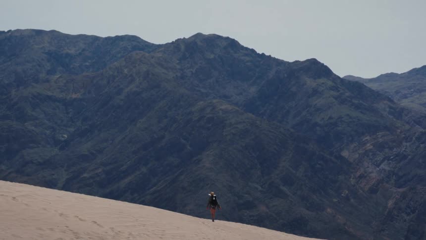 A hiker with a backpack walks across a vast sand dune towards a range of dark, rugged mountains
