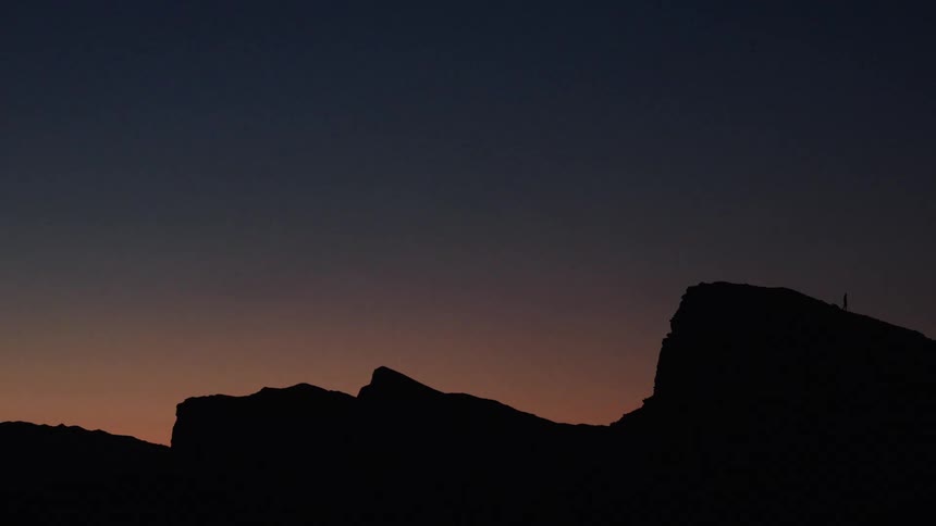 A tiny figure of a person is silhouetted against the vast twilight sky as they walk along a mountain ridge