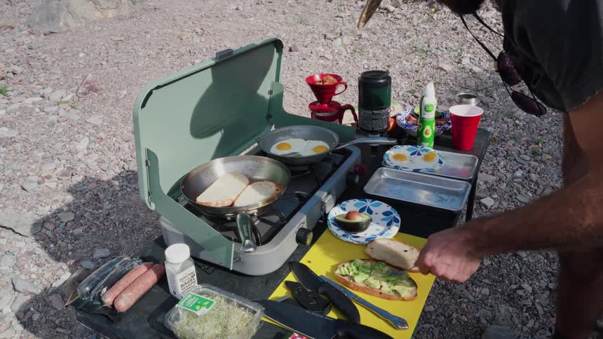 A man prepares a delicious breakfast of avocado toast and fried eggs on a portable camp stove while enjoying the outdoors
