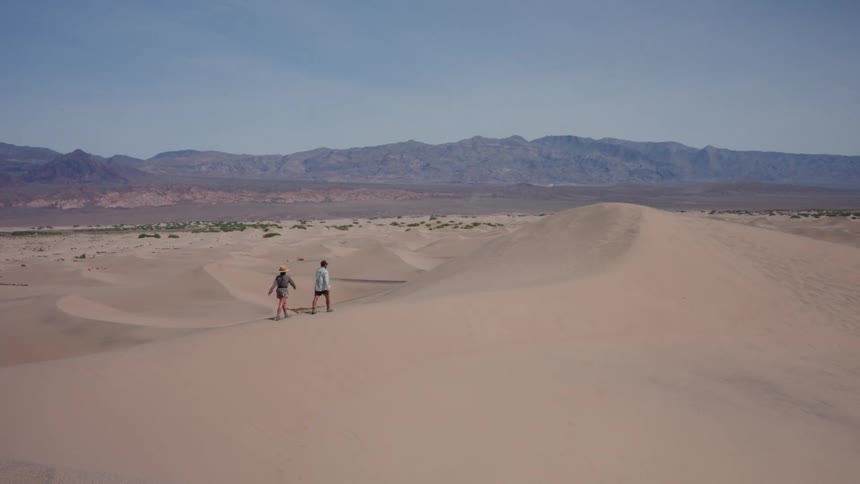 A man and a woman walk along the crest of a vast sand dune in the desert with mountains in the distance