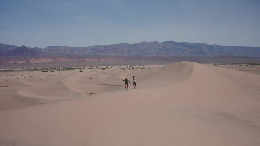 A couple joyfully hikes and runs in slow motion along the crest of a large sand dune in a vast desert valley
