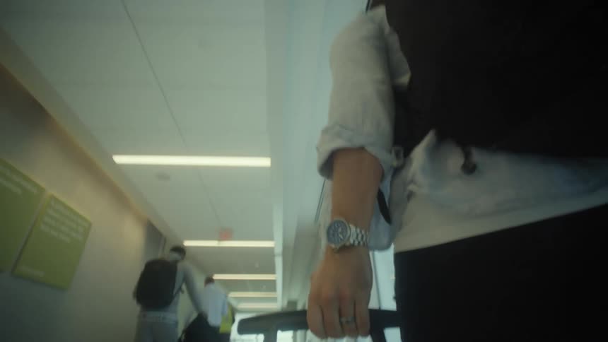 A low-angle follow shot of a person with a backpack pulling a rolling suitcase through an airport terminal hallway
