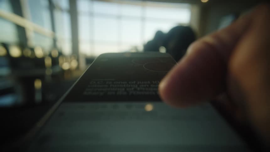 A dramatic macro close-up shot shows a thumb scrolling through a social media feed on a smartphone in a brightly lit airport terminal