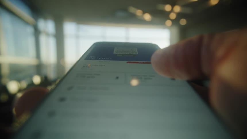 A traveler walks through a sunny airport terminal holding a smartphone displaying a digital boarding pass with a QR code