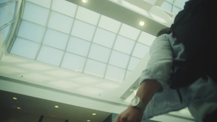 A low-angle shot follows a traveler with a backpack and suitcase walking through a sunlit airport terminal