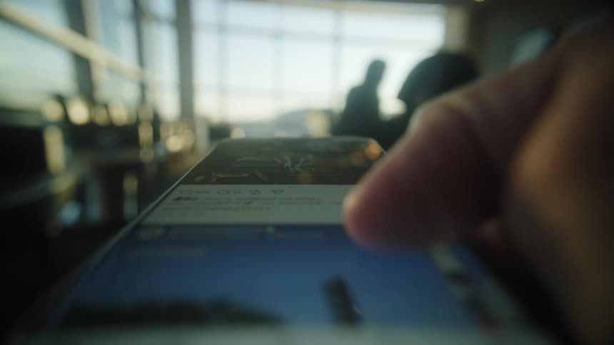 A person scrolls through their social media feed on a smartphone in an airport departure lounge, with the background blurred