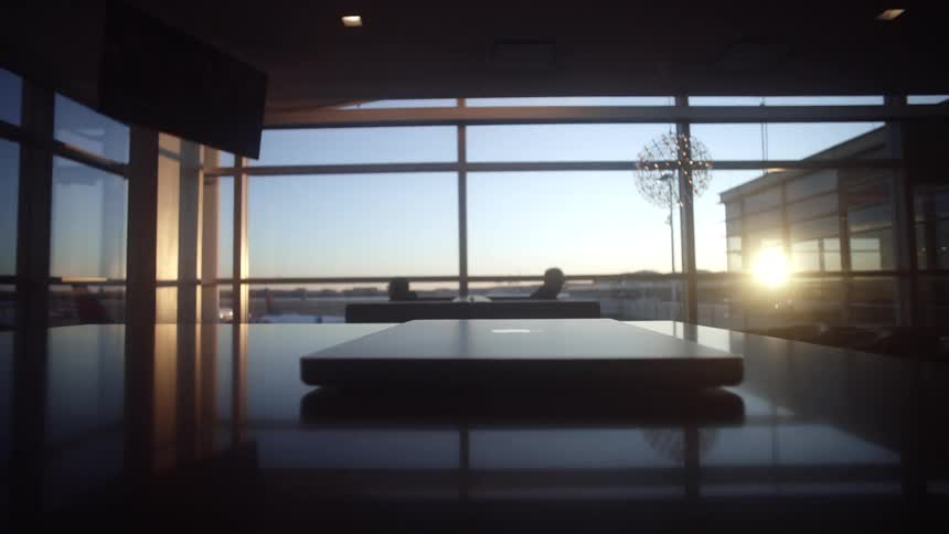 A silhouetted person finishes working and slowly closes their laptop on a table in an airport lounge at sunset