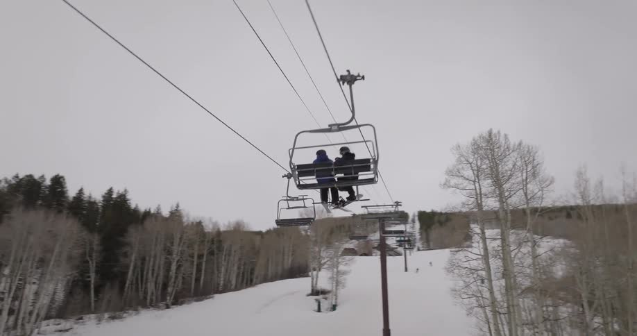 Ascending a snowy mountain on a chairlift, passing other skiers and snowboarders on an overcast winter day