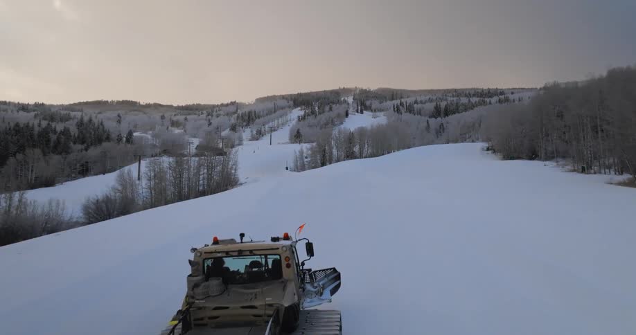 An aerial drone shot flies over a snowcat grooming a pristine ski slope at a mountain resort at sunset