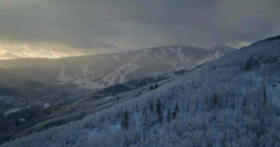 A majestic aerial view flies over a snow-covered forest toward a ski resort as sunlight breaks through the dark clouds