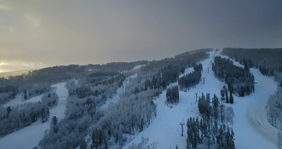 A slow aerial pullback reveals a vast, empty ski resort with frosted trees and fresh snow covering the slopes on an overcast winter day