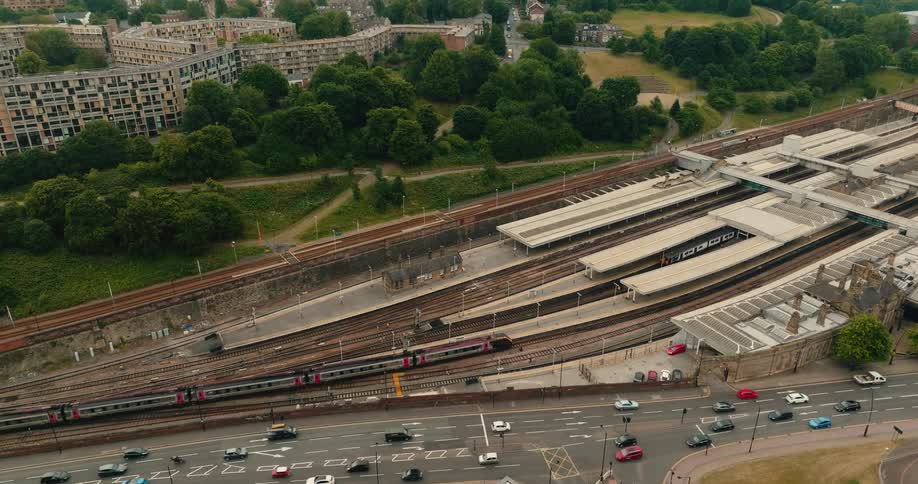 Aerial footage shows a passenger train arriving at a city station next to a road with traffic