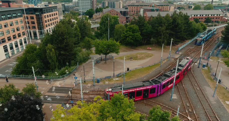 High angle aerial shot follows a modern red tram as it travels along curving tracks through a green urban space