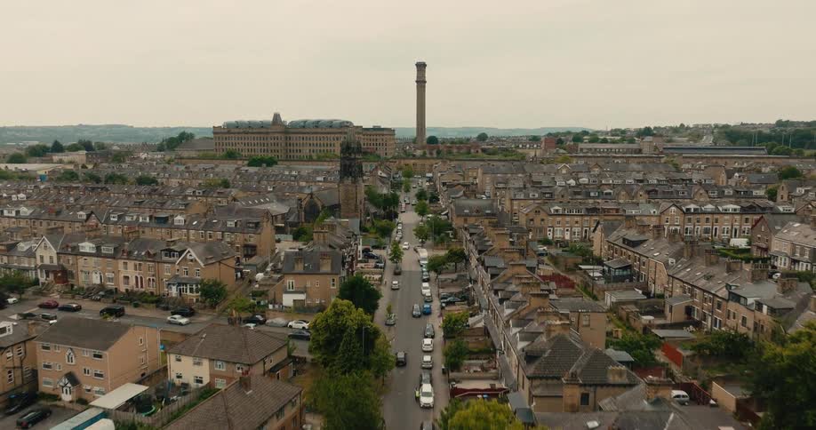 Drone slowly flies over a traditional English town revealing rows of terraced houses and a historic mill with a tall chimney on an overcast day