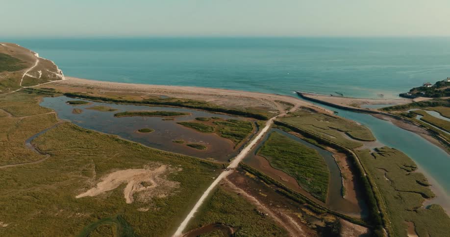 An aerial drone view pulls back to reveal a stunning coastal landscape where a river meanders through a lush green valley to meet the sea beside iconic white cliffs