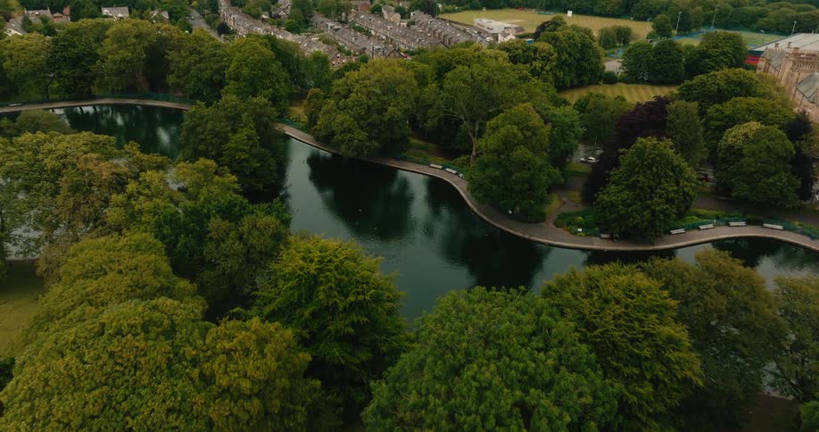 Aerial footage slowly tilts down over a beautiful park with a serpentine lake surrounded by lush green trees and a town in the background