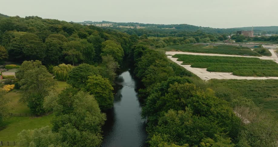 Slow aerial retreat over a dark river surrounded by dense green forests and rolling hills on an overcast day