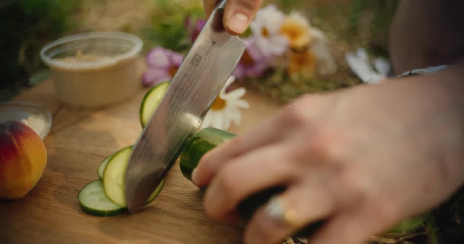 A close-up slow-motion shot shows hands slicing a fresh cucumber on a wooden cutting board for a beautiful outdoor picnic