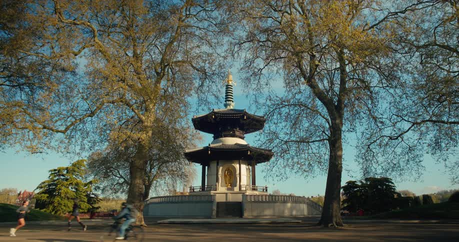 A wide, static shot shows the London Peace Pagoda in Battersea Park, framed by trees on a sunny day as people run and cycle past