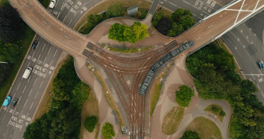 A top-down aerial view shows a tram crossing a unique three-way bridge over a complex railway junction as cars drive on adjacent roads