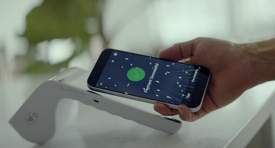 A hand holds a smartphone showing a 'Payment Successful' screen with animated confetti after completing a transaction on a contactless payment reader