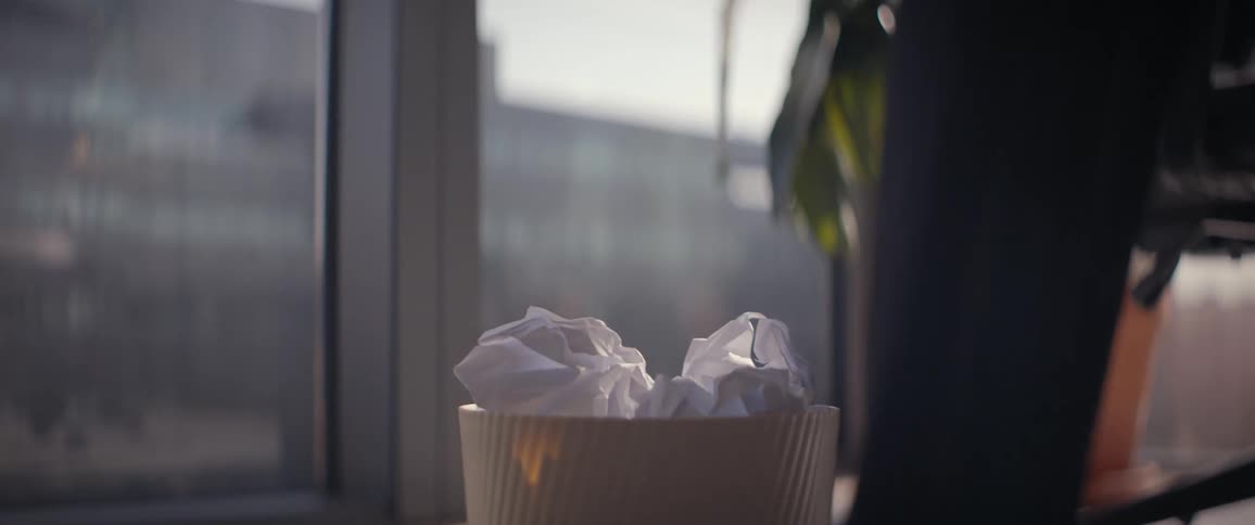 A crumpled paper ball is tossed into a full wastebasket in a sunlit office in slow motion
