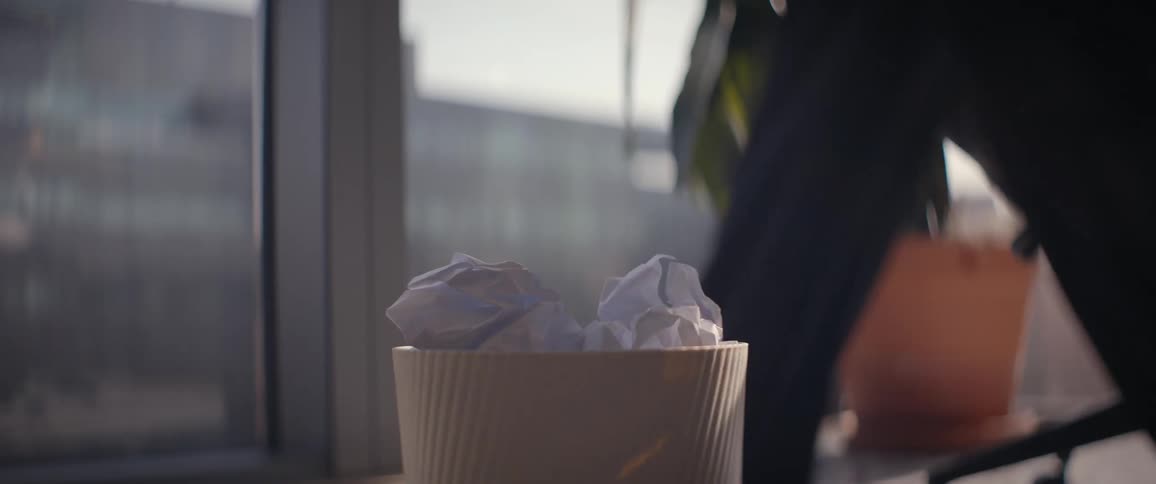 A close-up slow-motion shot of a person throwing a crumpled paper ball, missing the trash can in a sunny office