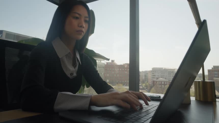 Slow push-in shot of a focused young Asian businesswoman typing on a laptop in a modern office with a city view