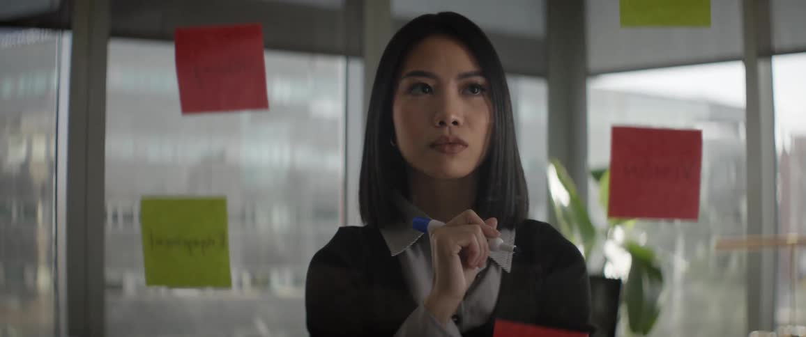 A focused young Asian businesswoman thoughtfully analyzes sticky notes on a glass wall during a brainstorming session in a modern office