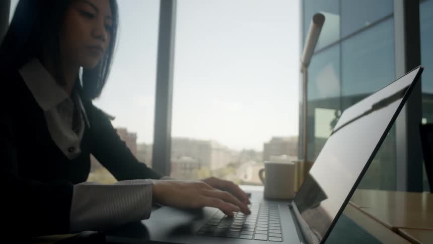 A close-up shot of a keyboard pulls back to reveal a young Asian businesswoman typing on a laptop in a modern office overlooking the city
