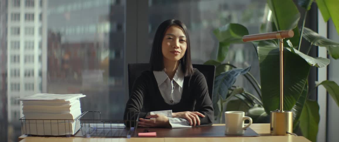 A thoughtful Asian businesswoman sits at her desk in a sunlit modern office and turns to look confidently at the camera