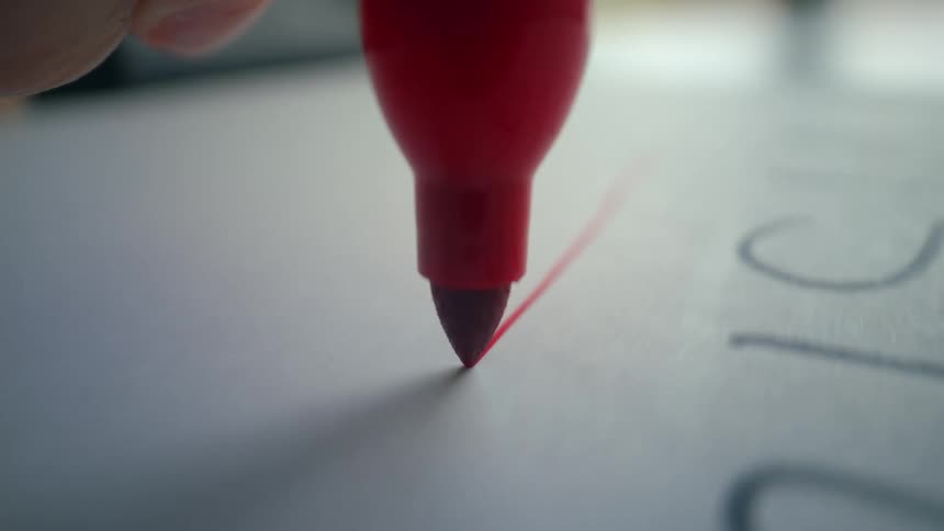 A close-up, slow-motion shot shows the tip of a red marker drawing a line on a white piece of paper