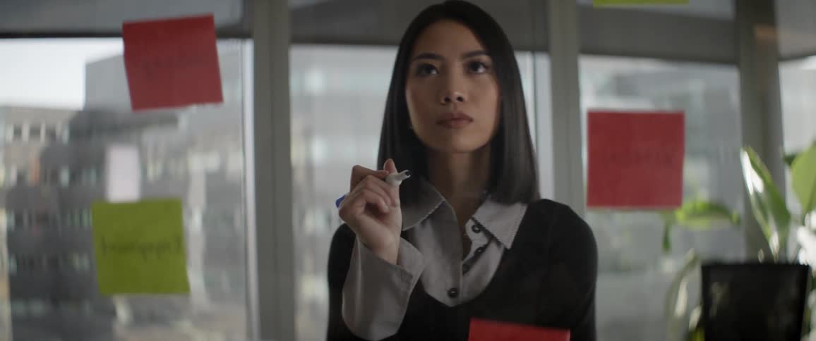 A focused young Asian businesswoman thoughtfully approaches a glass wall with sticky notes to brainstorm ideas in a modern office