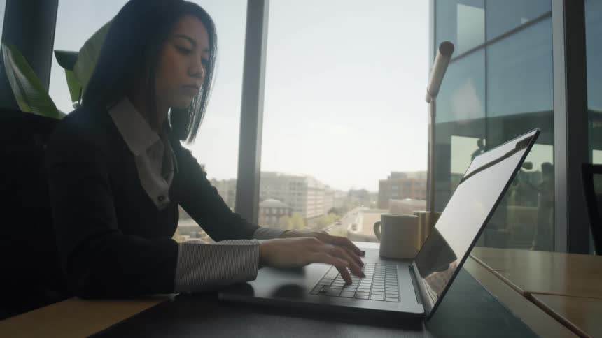 A professional Asian woman works on her laptop in a bright, modern office with a city view from a large window
