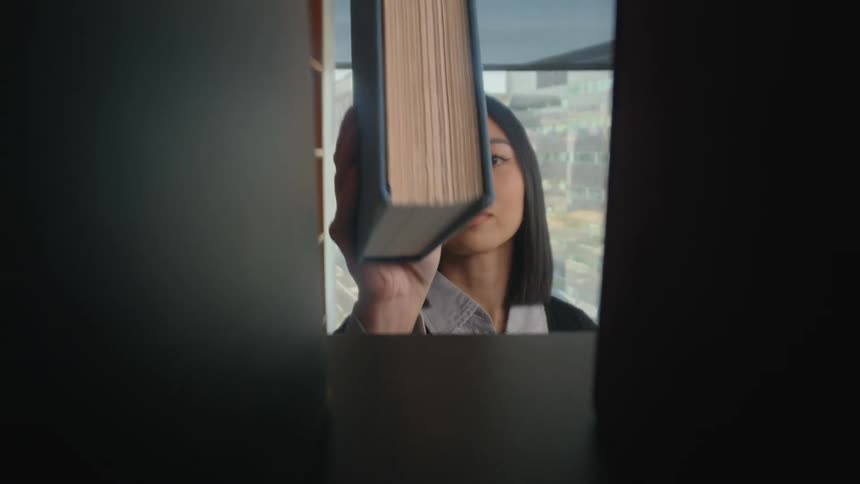 A woman places a large book onto a wooden bookshelf in a modern office, obscuring the view from within the shelf