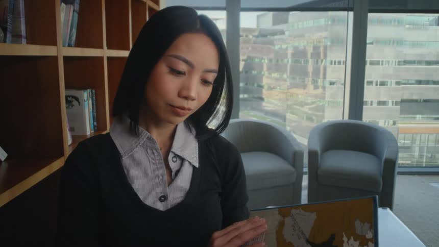 A young Asian woman takes an old book from a shelf in a modern city office and begins to read it