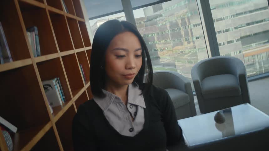 A young Asian woman pulls a large book from a shelf in a modern city library and begins to read it
