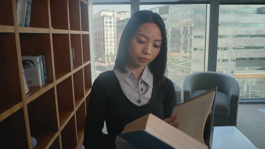 View from inside a bookshelf as a young Asian woman pulls out a large book in a modern office library with a city view