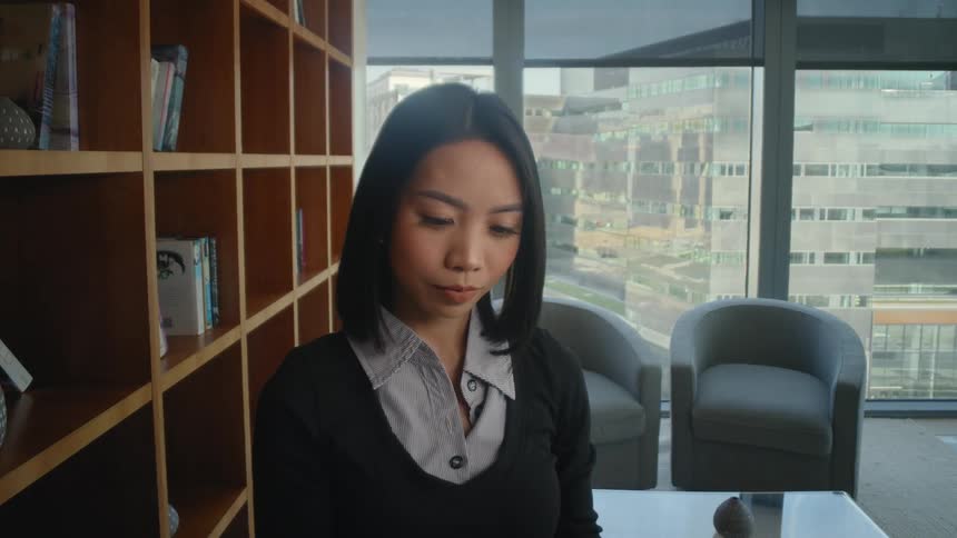 A young Asian businesswoman takes a book from a bookshelf in a modern high-rise office with a city view