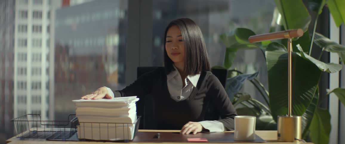 Pleased Asian businesswoman finishes signing a document, adds it to a large stack of paperwork, and leans back in her chair feeling accomplished
