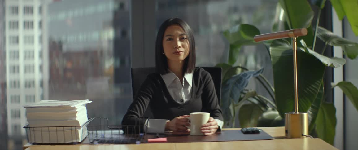 A thoughtful Asian businesswoman takes a moment to look out her office window before turning to look at the camera with a confident expression