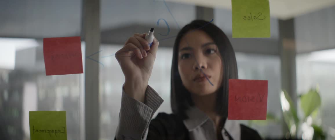 A focused young Asian businesswoman writes on a glass wall with a marker, connecting ideas from sticky notes during a brainstorming session in a modern office