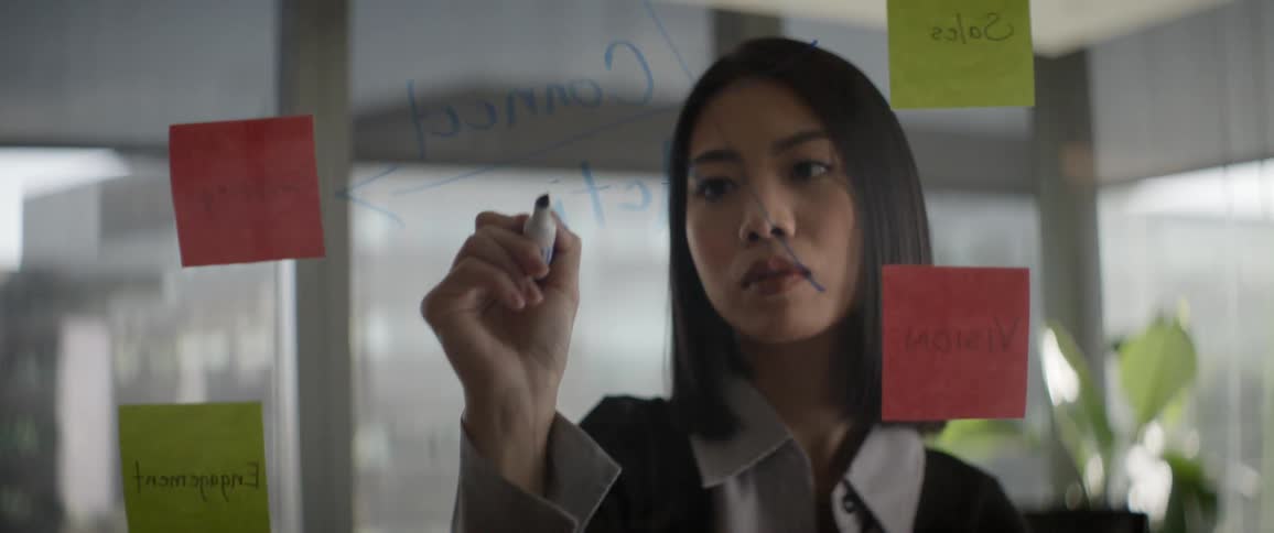 A focused Asian businesswoman brainstorms a strategic plan by writing on a glass wall with sticky notes in a modern office