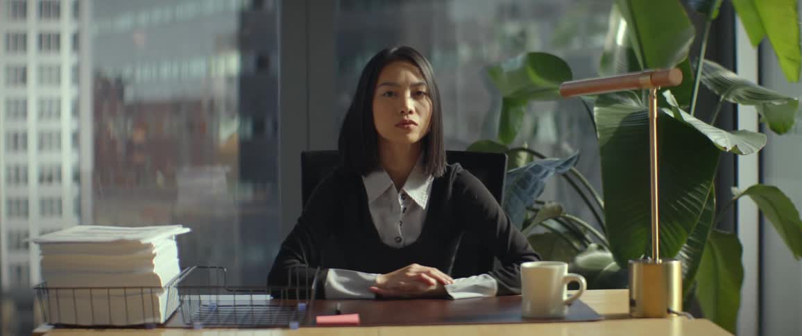 A thoughtful young businesswoman gazes out her city office window before turning to look directly and seriously at the camera