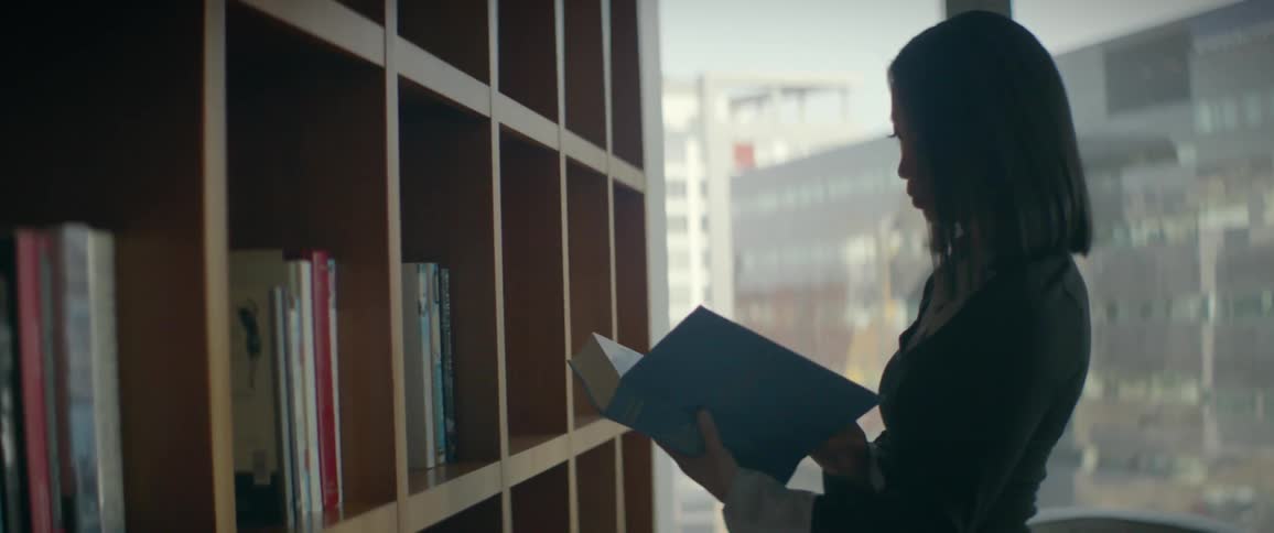 A professional Asian woman selects a book from a bookshelf in a modern office and begins to read it by the window