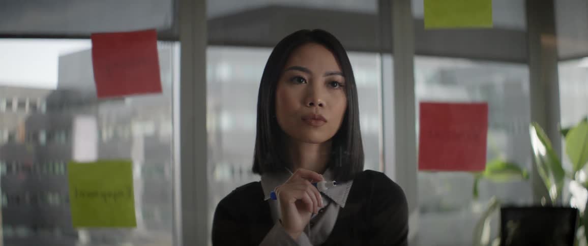 A focused Asian businesswoman in a modern office thoughtfully strategizes while looking at a glass wall covered in colorful sticky notes