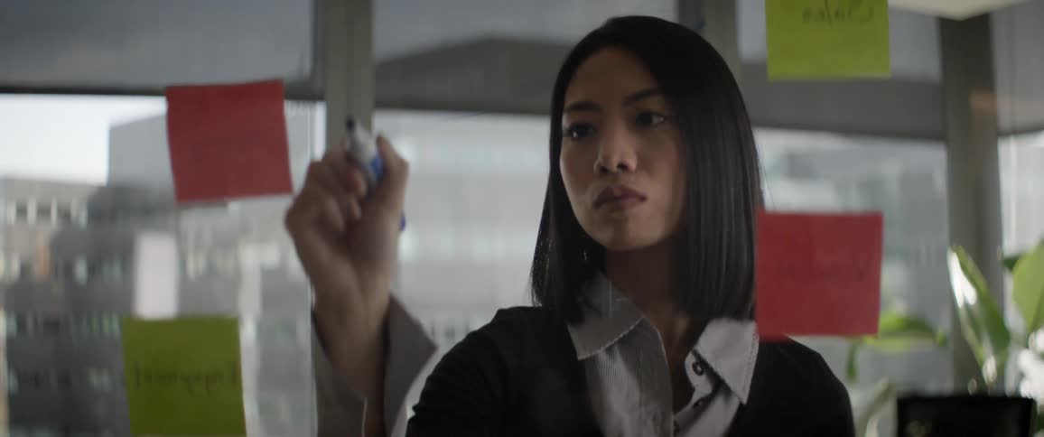 A focused young Asian businesswoman strategizes and brainstorms, writing on a glass wall with sticky notes in a modern office